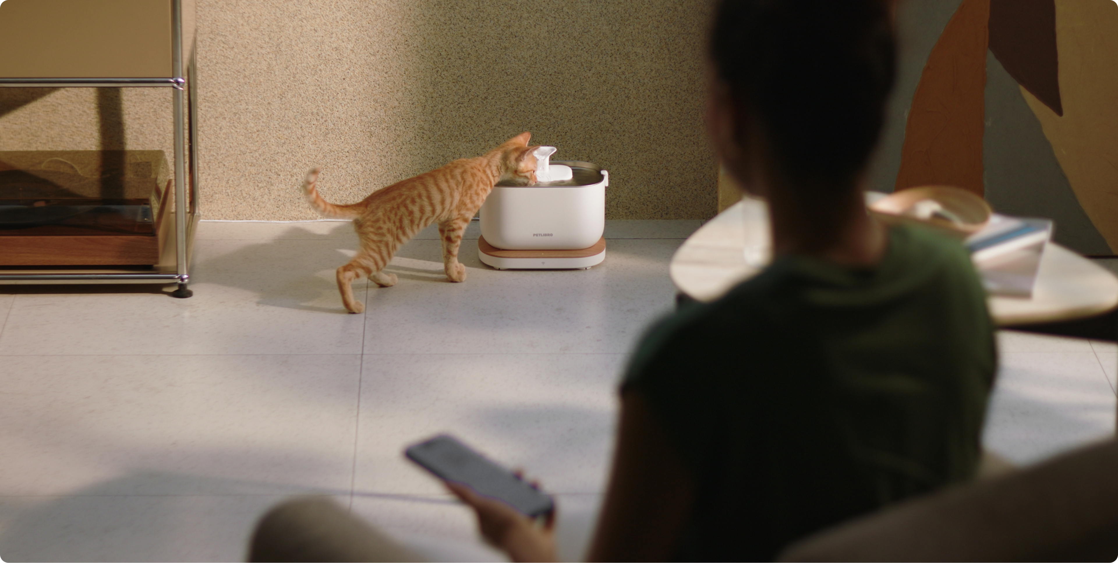 Petlibro automatic pet water fountain in white with wooden base, dispensing water for a drinking orange tabby cat in a modern home setting